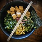 Noodle bowl with tofu, seaweed, greens, corn, and chopsticks on a dark wooden table.