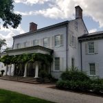 Historic white farmhouse with columns and mature trees under a cloudy sky.