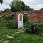 Brick wall with white arched gate, lush greenery and garden path under cloudy sky.