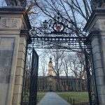 Ornate wrought iron gate leading to a grand, historic building through bare trees.