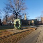 Green abstract sculpture in a park setting with historic buildings under a blue sky.