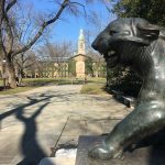 Bronze lion statue in foreground with a university building and trees under a blue sky.