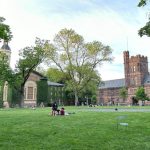 University campus quad with historic buildings, green lawn, and trees under a bright sky.