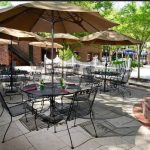 Outdoor patio dining area with tables, chairs, and large umbrellas under green trees.