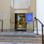 Entrance to a classical building with columns and an "Our Sanctuary is open" sign.