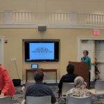 People gather in a bright hall watching a presentation on a screen.
