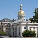 Capitol dome with golden top against a blue sky, surrounded by classical government buildings.