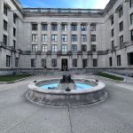 Neoclassical building facade with a central fountain and bronze statue in the courtyard.