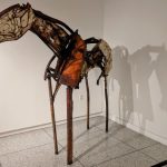 Deconstructed, skeletal horse sculpture in a white gallery space with prominent shadow.