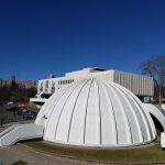 Large white geodesic dome structure in front of a modern building under a clear blue sky.
