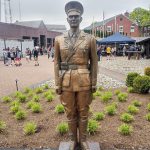 Bronze statue of a soldier stands in a park setting with buildings and trees in the background.