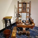 Antique electric chair display with wooden structure and warning signs on a blue rug.