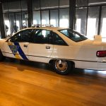 White and blue State Police car parked indoors with large windows in the background.