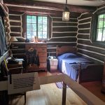 Rustic cabin interior with log walls, wooden furniture, and multiple windows.