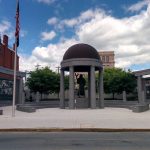 Bronze statue under a domed monument in a plaza with surrounding brick buildings.