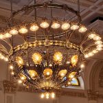 Ornate, large brass chandelier illuminating a grand interior hall with detailed architectural molding.