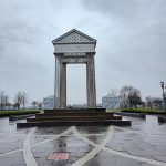 Modern, geometric pavilion structure under a gray, overcast sky in a public plaza.