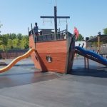 Wooden pirate ship playground with two slides on a paved outdoor area.