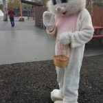 Costumed white rabbit character holding a basket stands on dark ground outdoors.