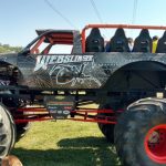 Monster truck with riders on top, parked outdoors in a grassy field under sunny skies.