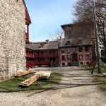 Stone castle wall beside historic buildings and dirt path under a bright sky.