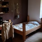 Rustic bedroom interior with wooden bed, shelves of pottery, and natural light from a window.