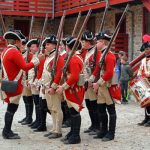 Musketeers in historical uniform stand outdoors near a stone building.