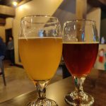 Two glasses of amber and dark beer on a wooden bar counter indoors.