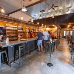 Rustic bar interior with long counter, beer taps, and patrons standing.
