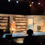 Stage set with shelves and a doorway under bright spotlights for a performance.