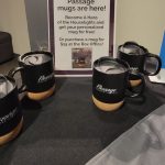 Assorted Passage mugs displayed near a sign promoting personalized mug availability.