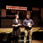 Two actors on a stage with a backdrop of framed artwork and a checkered floor.