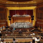 Ornate theater interior with a stage, orchestra seating, and audience members seated.