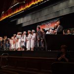 Graduation ceremony on stage with students in caps and gowns, speakers addressing the audience.