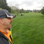 Man watches golfers on a lush green golf course under an overcast sky.