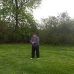 Golfer in dark attire stands on a green lawn with trees and bushes in the background.