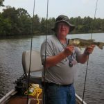 Man with hat holds a large fish while fishing from a boat on calm water.