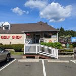 Golf shop building with porch and white railing, parking lot in front under blue sky.