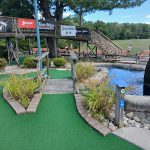 Putting green with wooden bridge and water feature, featuring golf equipment sponsors.