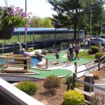 Park scene with a pond, playground area, and large trees under a bright sky.