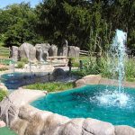 Outdoor water park scene with a large fountain, rock features, and lush green trees.