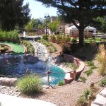 Tiered rock waterfall and pond landscaping in a sunny, lush garden setting.