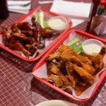 Two red trays filled with glazed chicken wings and dipping sauces on a checkered tablecloth.