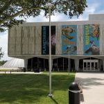 Modern building facade with "NJ State Museum" signage, set on a green lawn under trees.