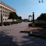 Neoclassical building and colonnade viewed down a wide, tree-lined walkway toward a distant archway.