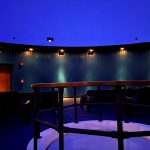 Dark theater interior with tiered seating and a curved stage area under blue lighting.