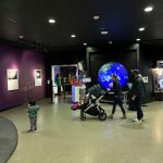 Family interacts with a large blue planet display in a modern, dark-themed exhibit hall.