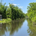 Calm waterway reflecting green trees under a bright blue sky.