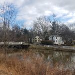 Rural scene with houses, trees, and a pond under a cloudy sky.