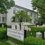 White historic church with surrounding cemetery and mature trees under an overcast sky.